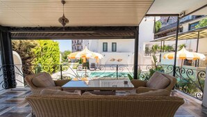 Un área de estar al aire libre cubierta con mobiliario de mimbre y sombrillas, con vistas a una piscina y edificios.