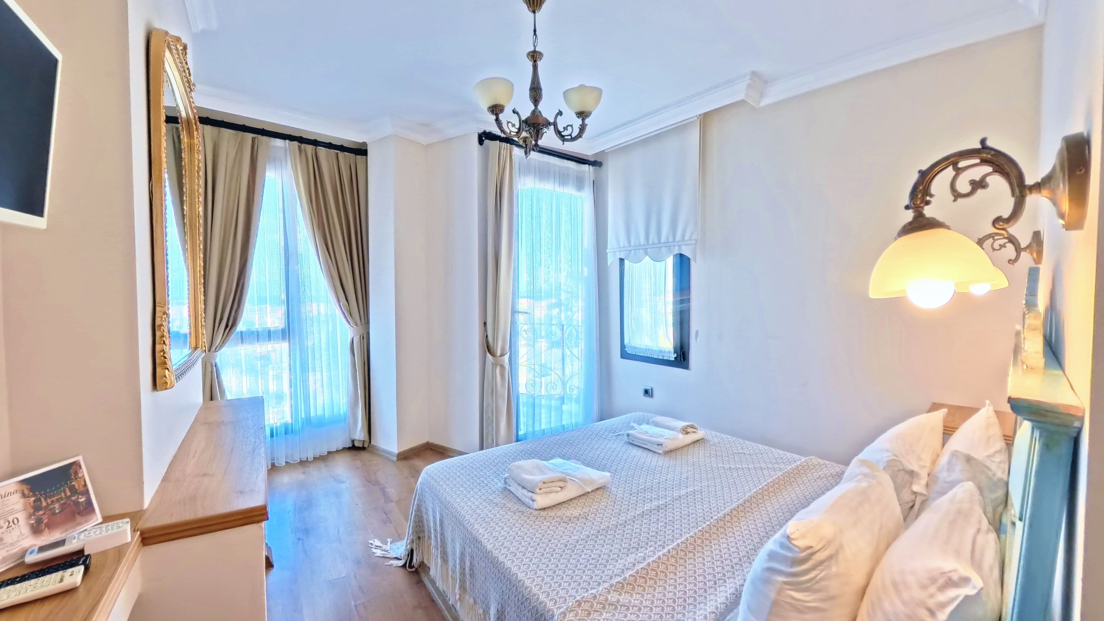 Una habitación de hotel con cama, candelabro, ventana con cortinas y lámpara de noche.