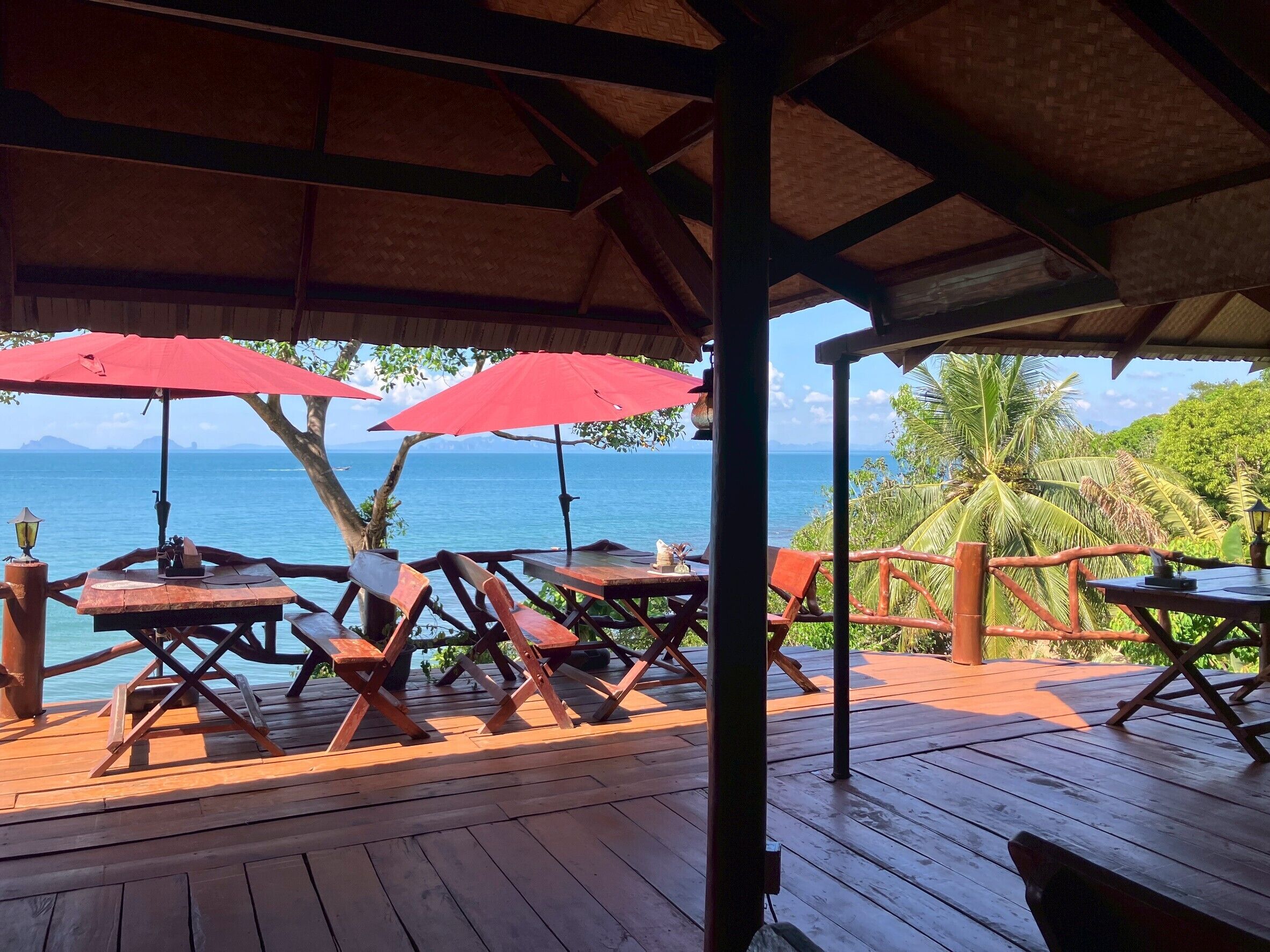 A wooden deck with tables and chairs, red umbrellas, and a view of the ocean.