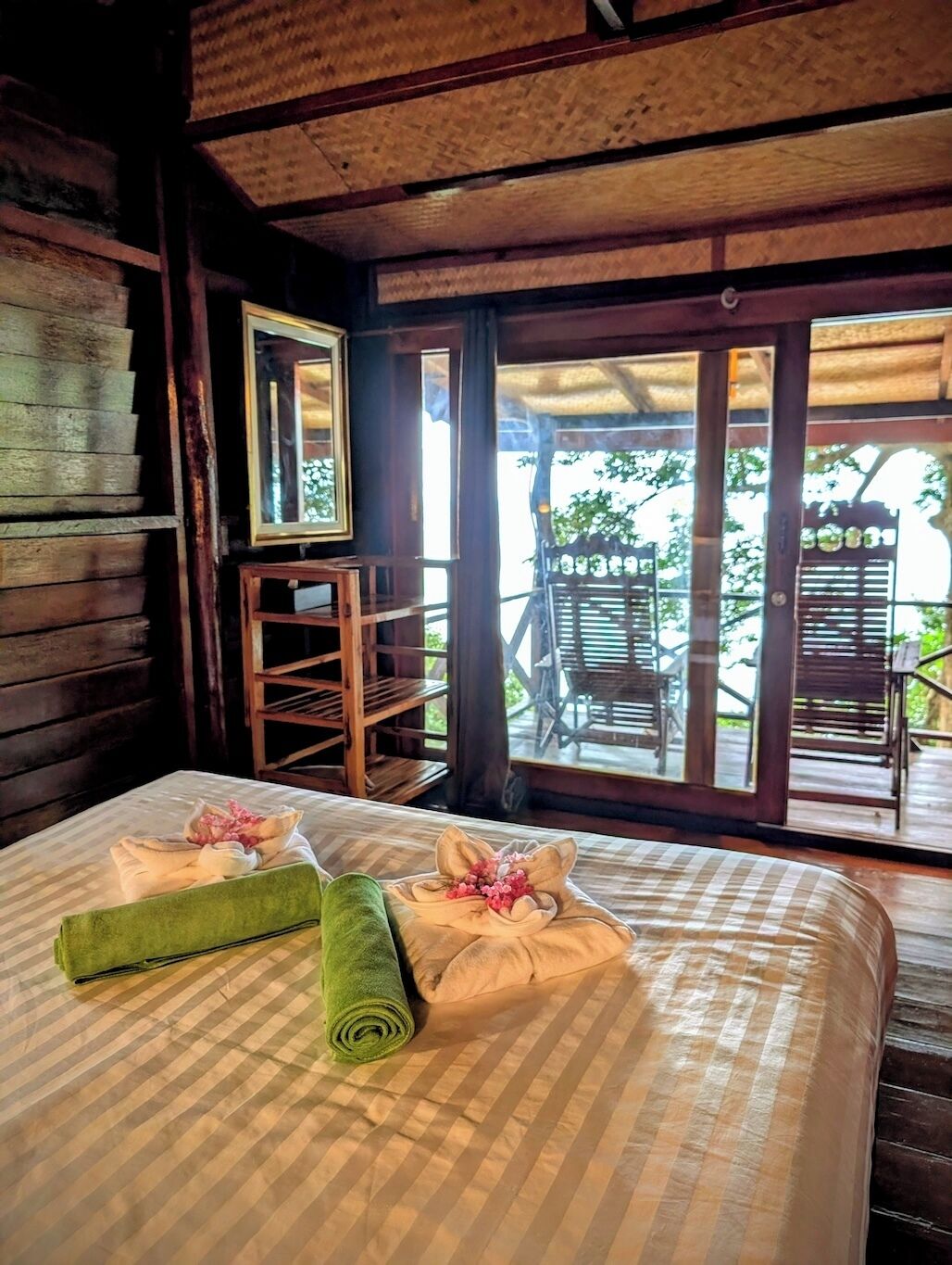 A wooden cabin room with a bed, a mirror, and a view of an outdoor patio with chairs.