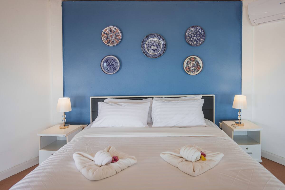 Un lit bien fait, avec du linge de lit blanc, deux tables de chevet avec des lampes, et un mur d’accent bleu orné d’assiettes décoratives.