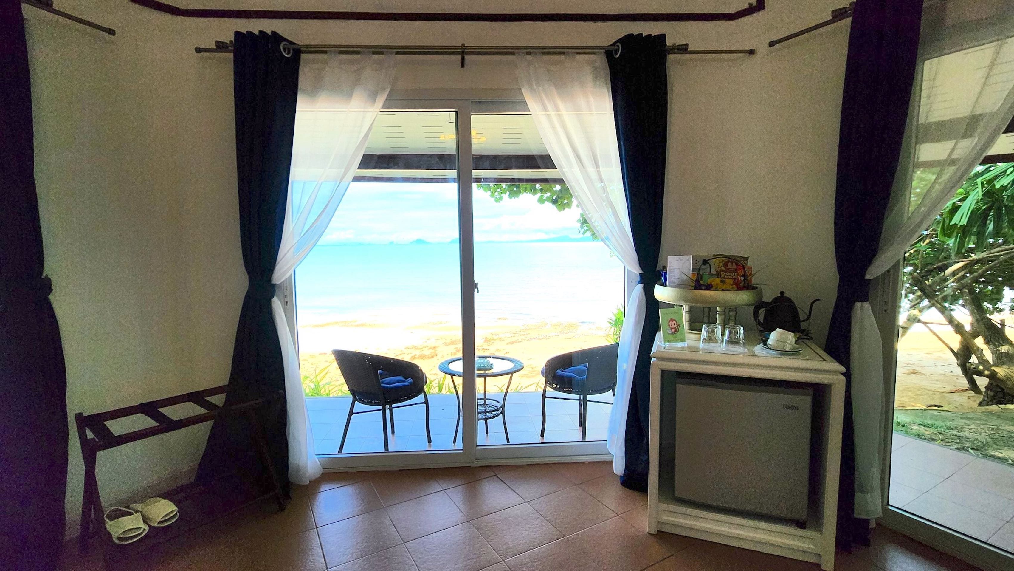A room with a view of the beach, two chairs, a small table, and a cabinet with items on it.