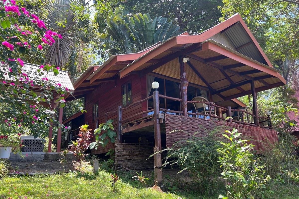 Une maison en bois au style rustique, dotée d’un balcon et entourée d’une végétation luxuriante et de plantes en fleurs.