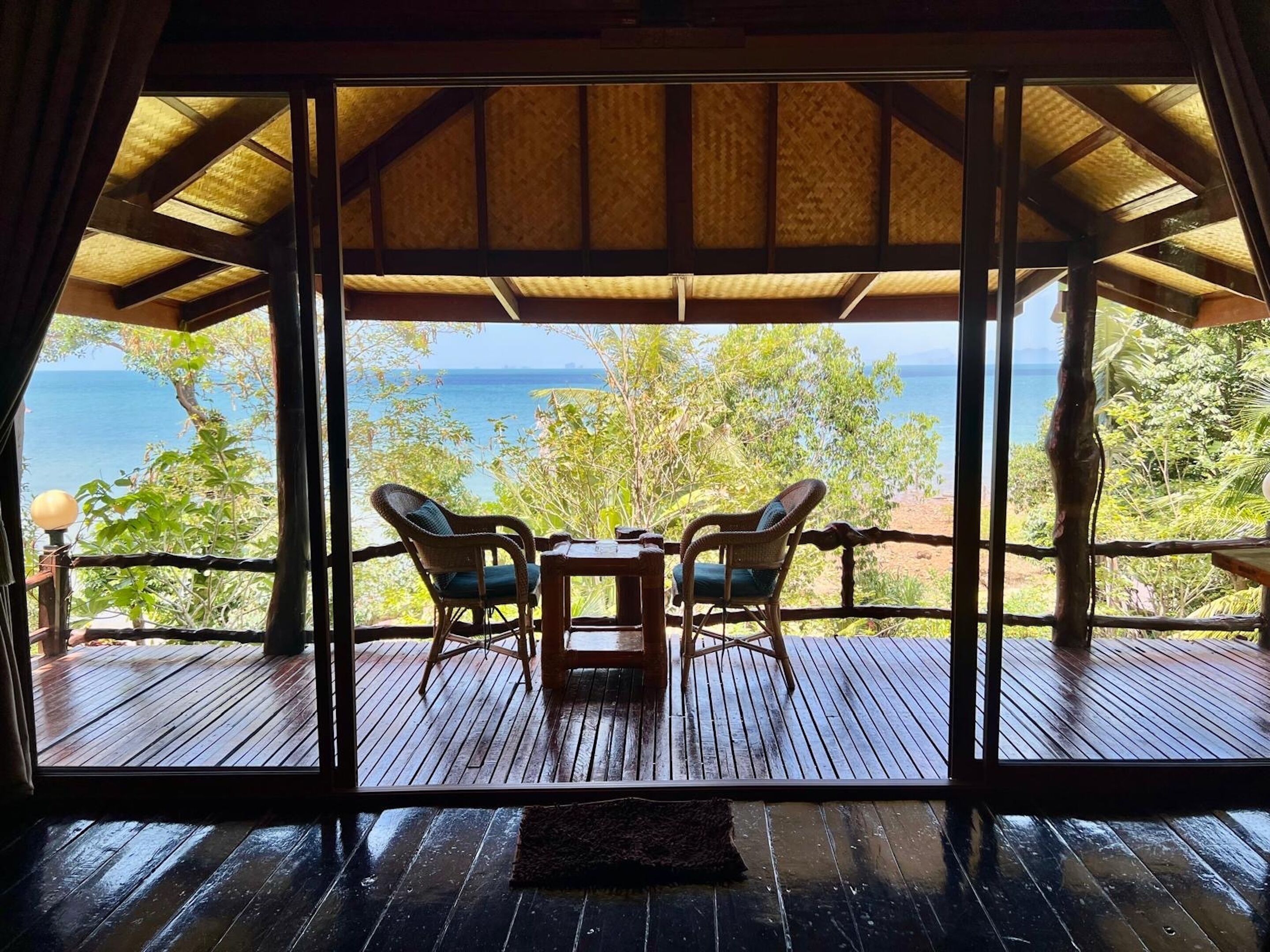 A wooden deck with two wicker chairs and a table, overlooking a beach and the ocean.