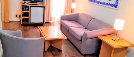 Un salon avec un canapé gris, une table basse en bois, un fauteuil gris, un téléviseur posé sur un meuble et un petit réfrigérateur.