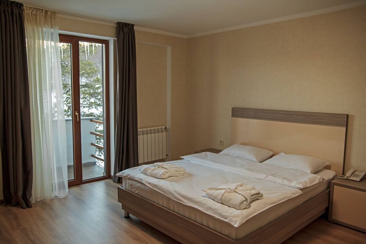 Habitación de hotel con una cama de madera, ropa de cama blanca, un teléfono junto a la cama y vistas a un balcón a través de una puerta de cristal.