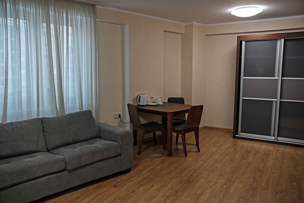 A living room with a grey sofa, a wooden dining table with four chairs, a coffee maker, and a sliding glass door.