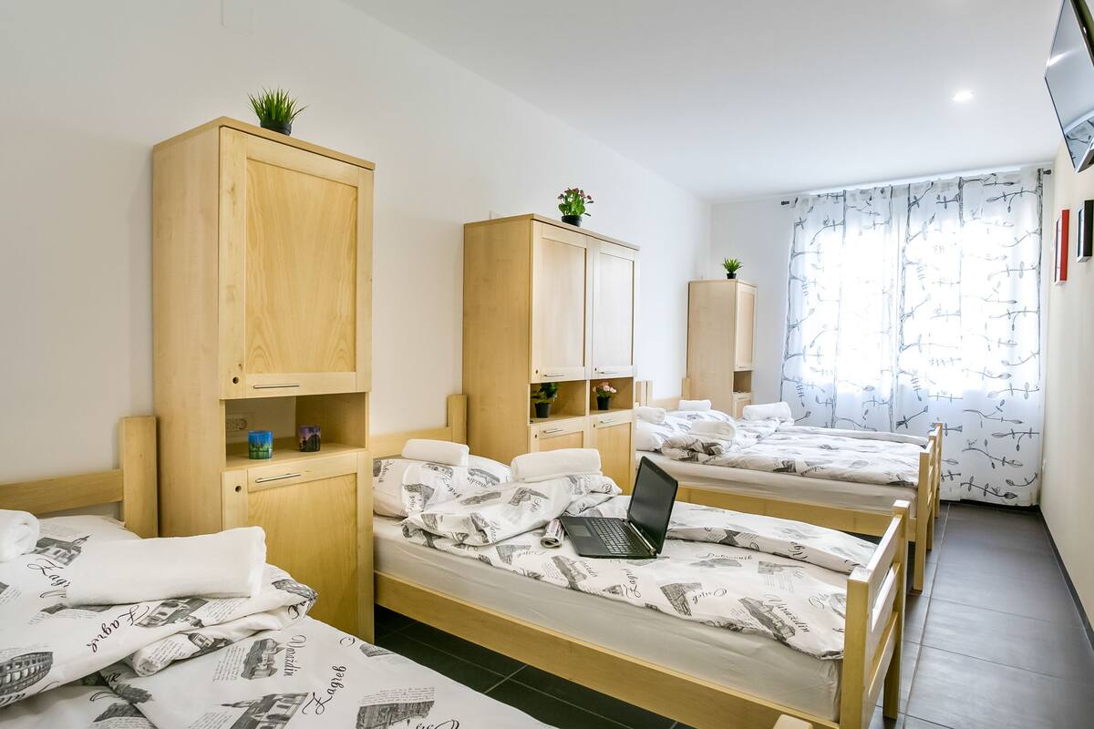 A dormitory room with three single beds, each with a laptop and a plant on top. There are wooden cabinets with small shelves, and a window with patterned curtains.