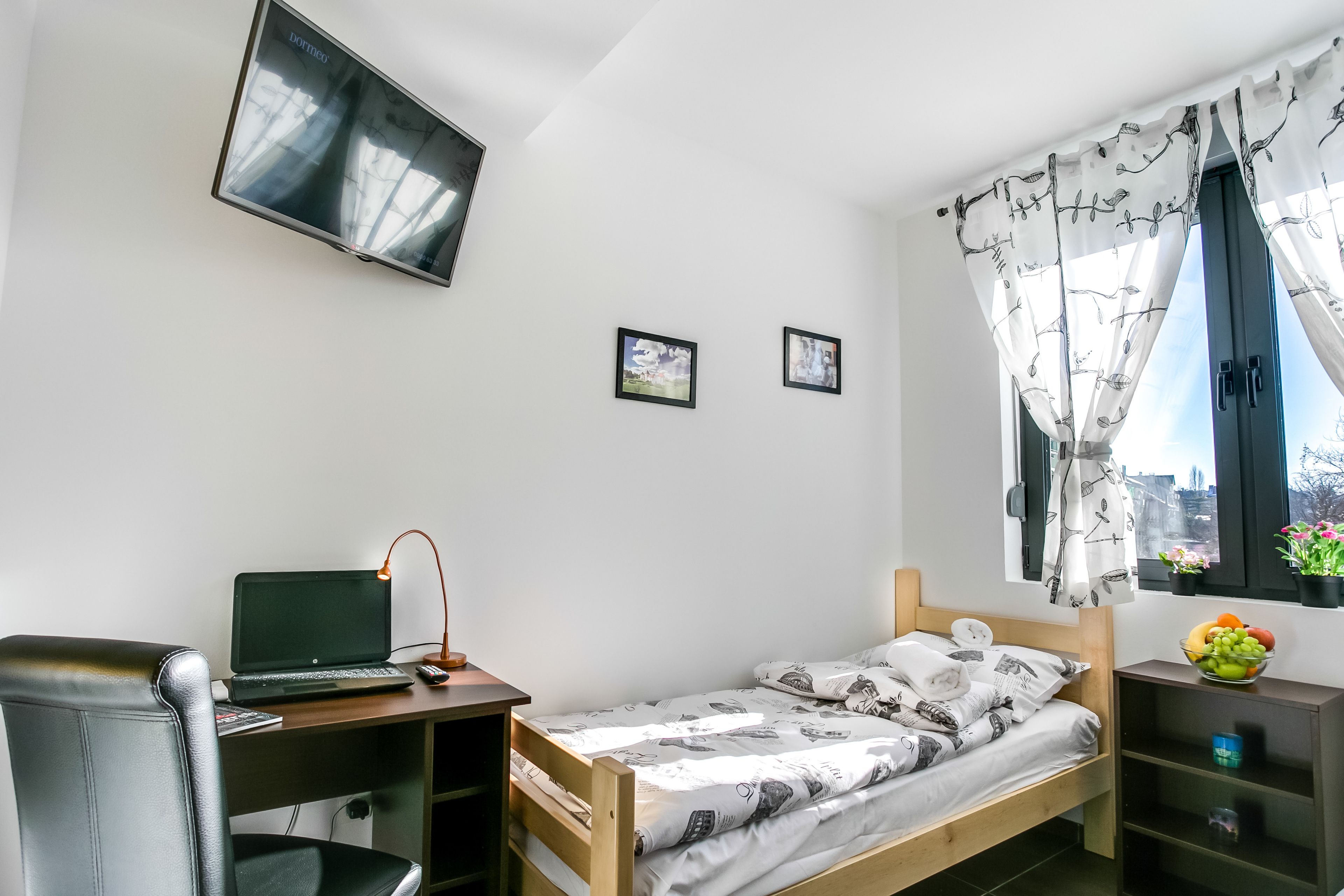 Una stanza piccola e ordinata con un letto, una scrivania con un portatile, una sedia e un televisore montato a parete.