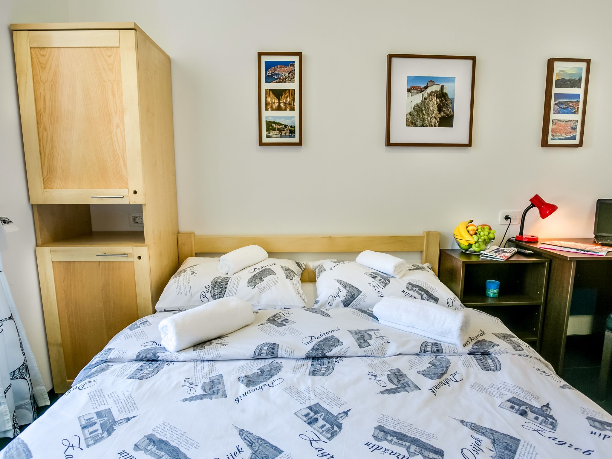 A neatly made bed with a patterned duvet, a wooden wardrobe, a desk with a lamp, and framed pictures on the wall.