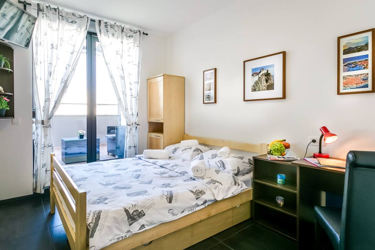 A neatly arranged bedroom with a bed, a desk, a chair, a small table, and a window with curtains.