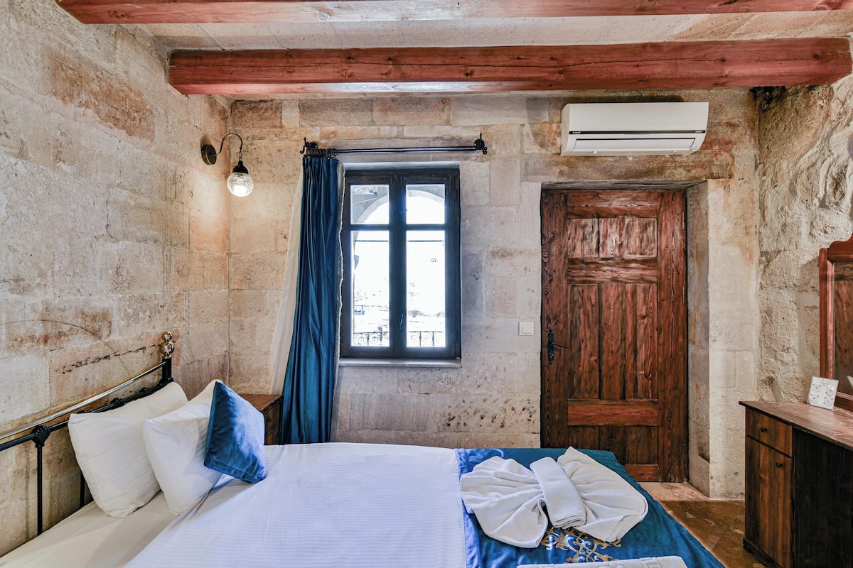 A bedroom with a bed, a window with curtains, a wooden door, and an air conditioning unit.