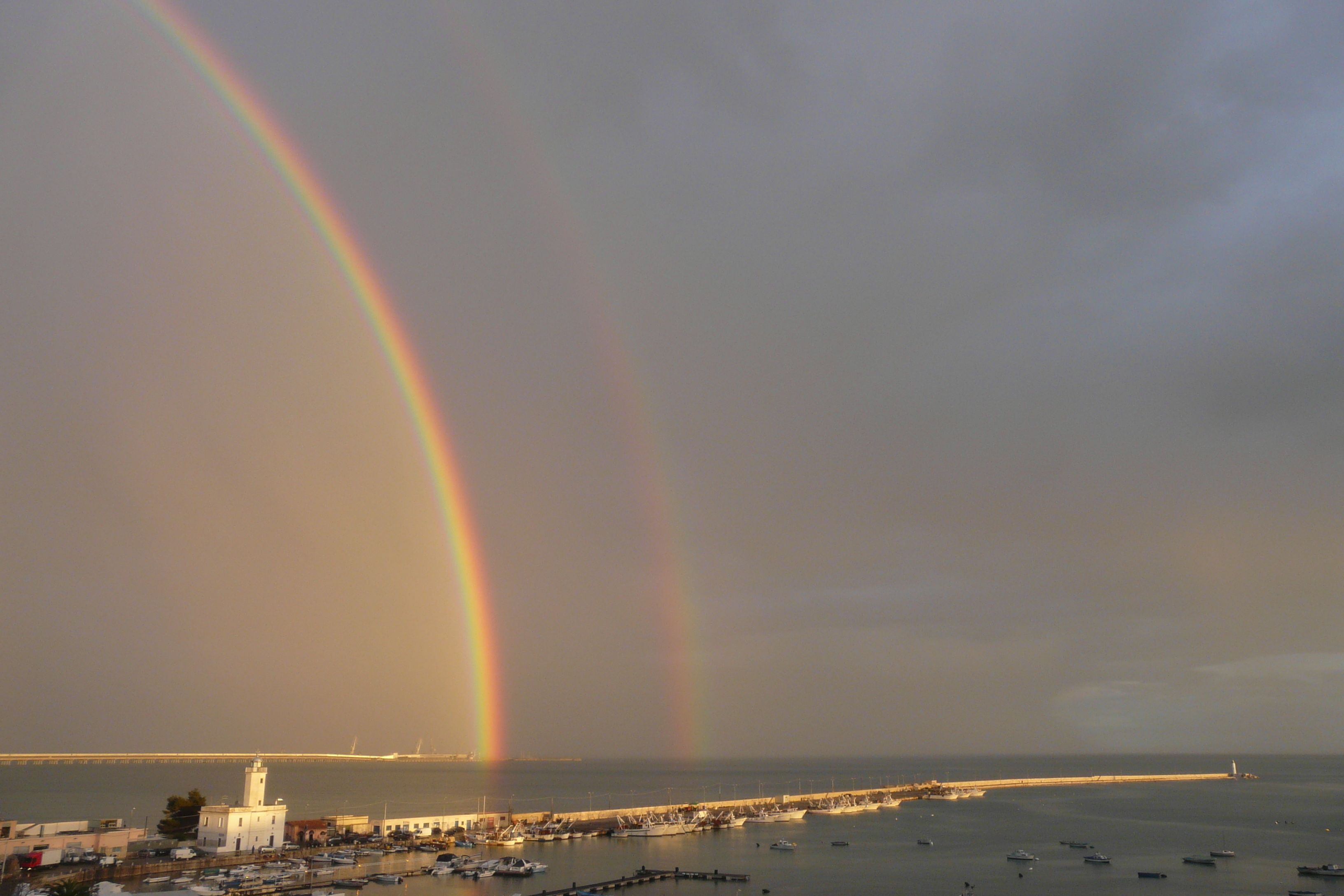 A coastal marina with boats docked, a lighthouse, and a rainbow in the sky.