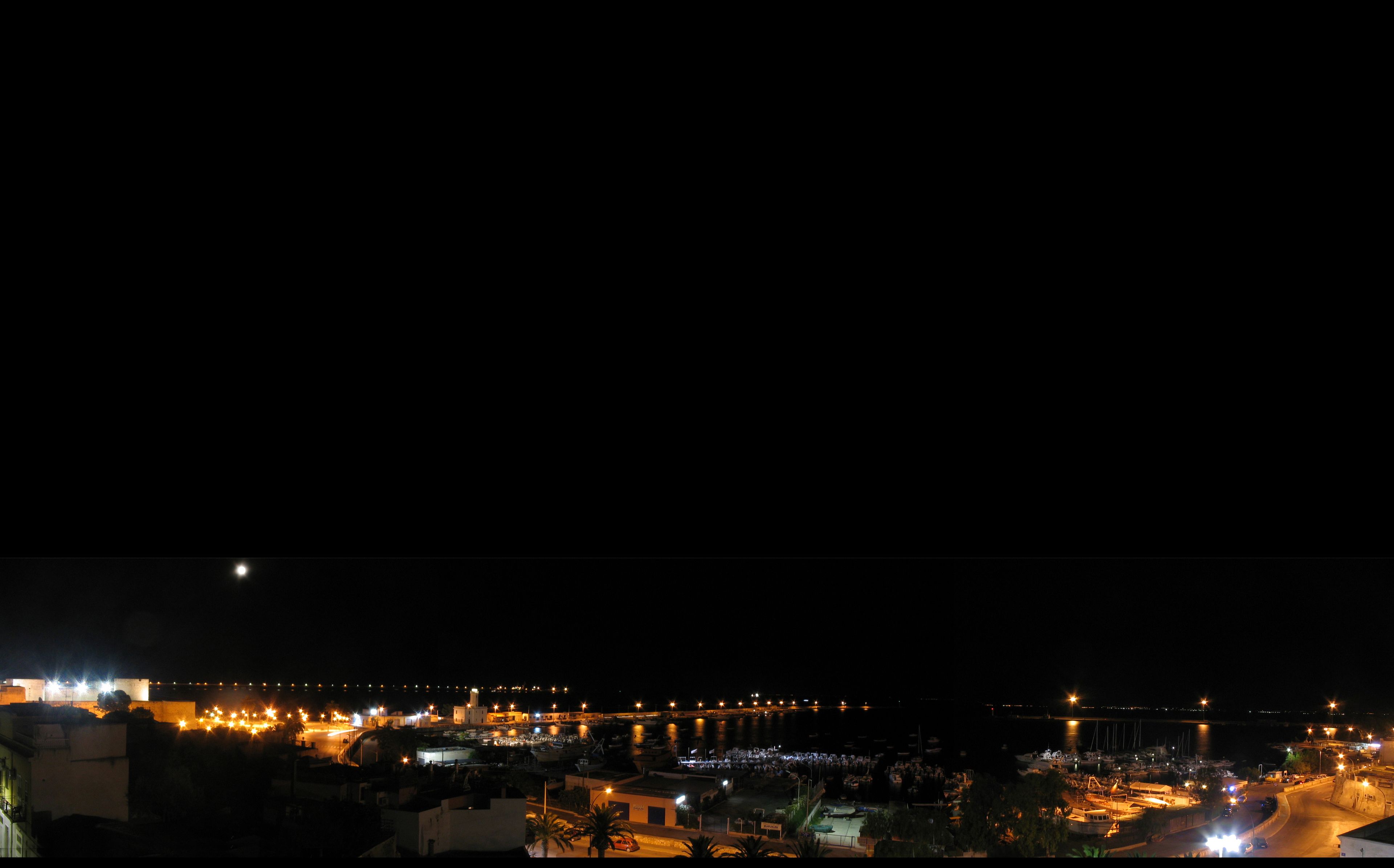 A nighttime view of a coastal town with illuminated buildings, a marina, and a bridge.