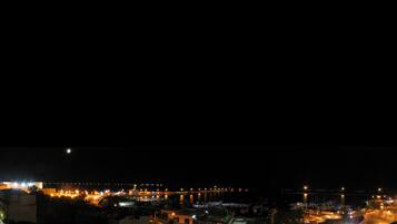 A nighttime view of a coastal town with illuminated buildings, a marina, and a bridge.