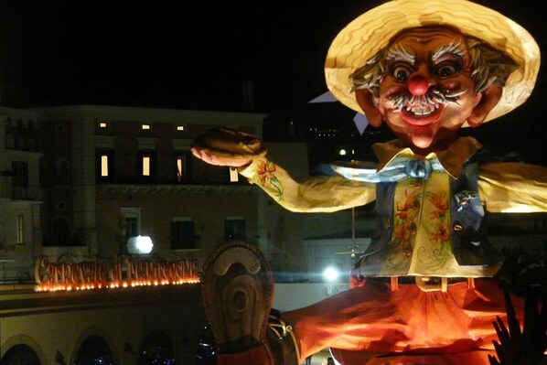 A large, illuminated sculpture of a character with a wide-brimmed hat and a colorful outfit, set against a building facade with illuminated windows. - La dolce vista (Manfredonia)