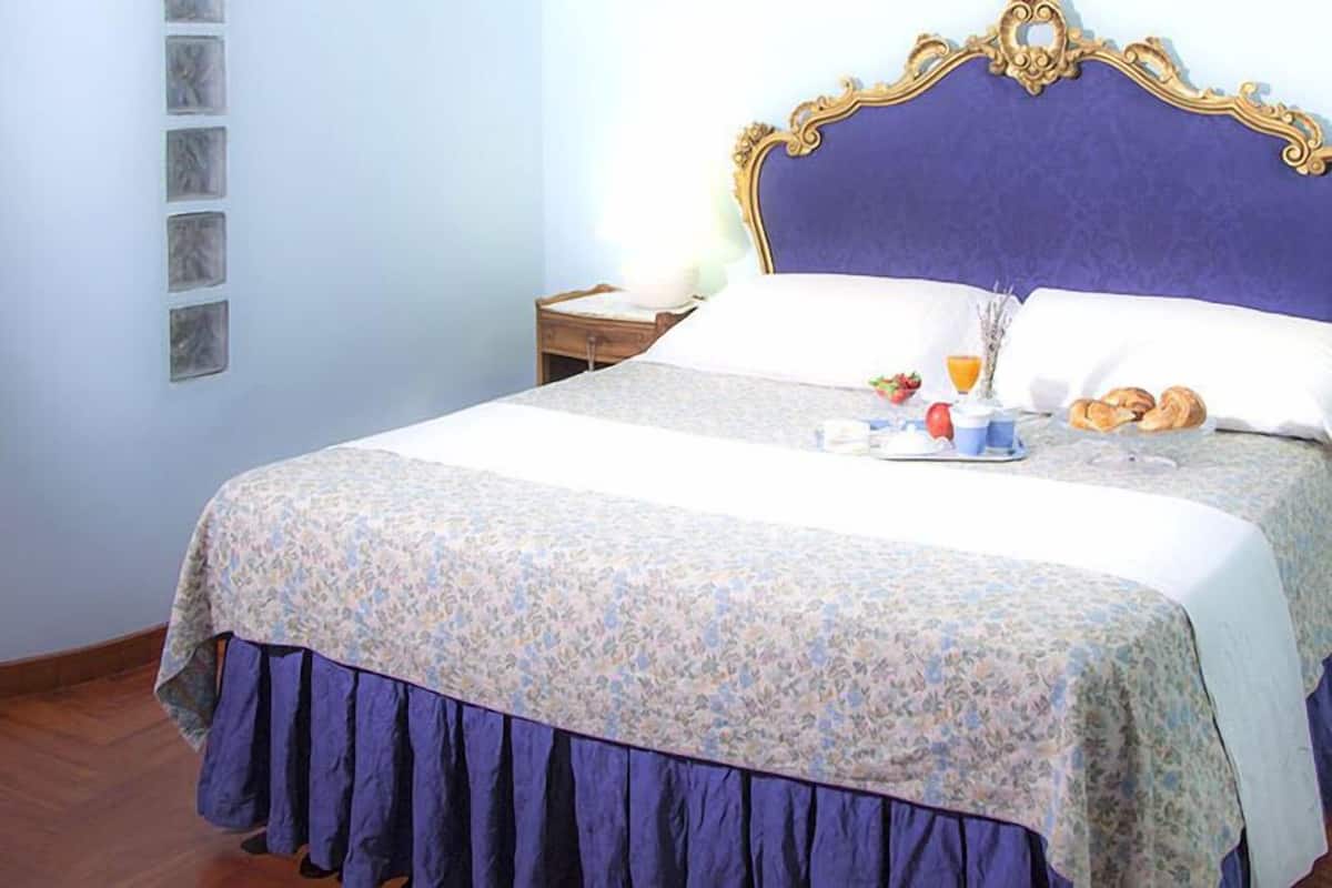 A neatly made bed with a floral bedspread and a purple skirt, a nightstand with a lamp, and a wall with a series of square mirrors.