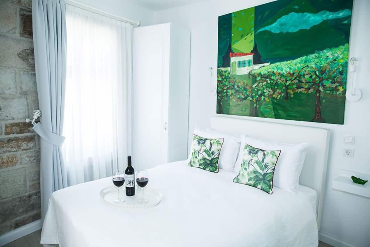 Una cama con colcha blanca, dos almohadas estampadas en verde, y una bandeja con botellas de vino y copas.