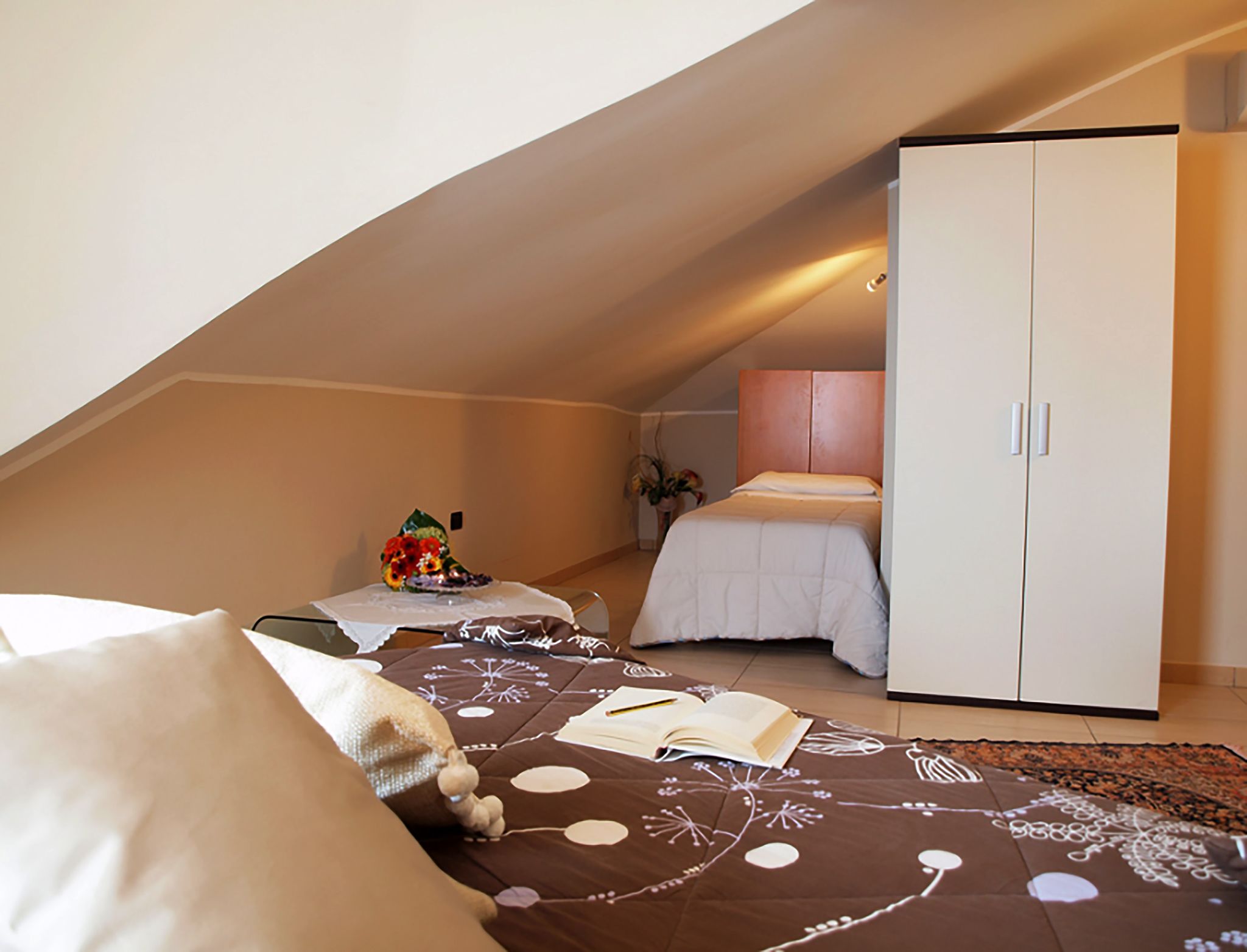 Une chambre avec un plafond en pente, un lit avec une tête de lit blanche, une table de chevet avec une plante et une armoire.