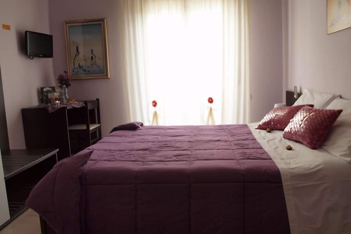 A bedroom with a large bed, a desk with a computer, a painting on the wall, and a window with curtains.