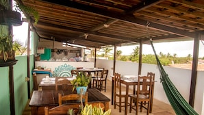 A covered patio with wooden tables and chairs, a kitchen area, and a view of palm trees. - Pousada A Casa é Sua (Jijoca de Jericoacoara)