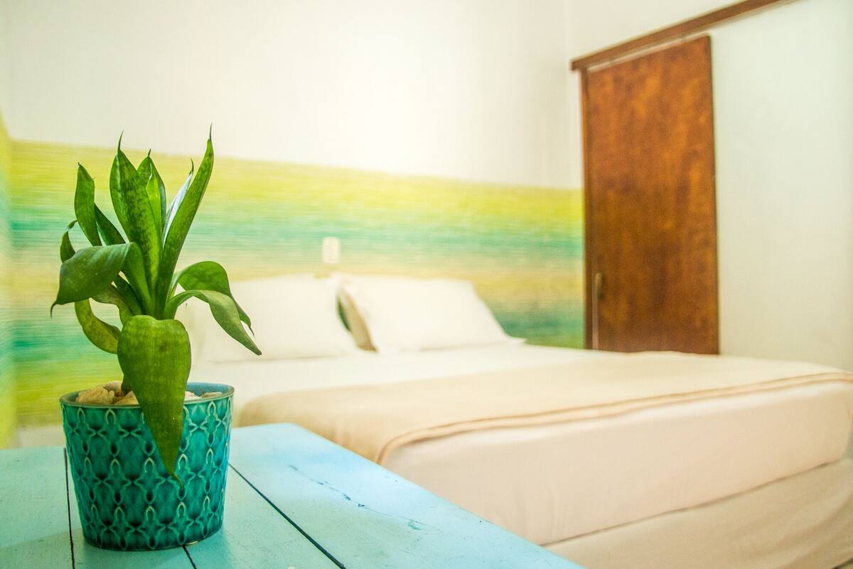Una cama con cabecera de madera azul, almohada blanca y planta en maceta verde sobre una mesa azul.
