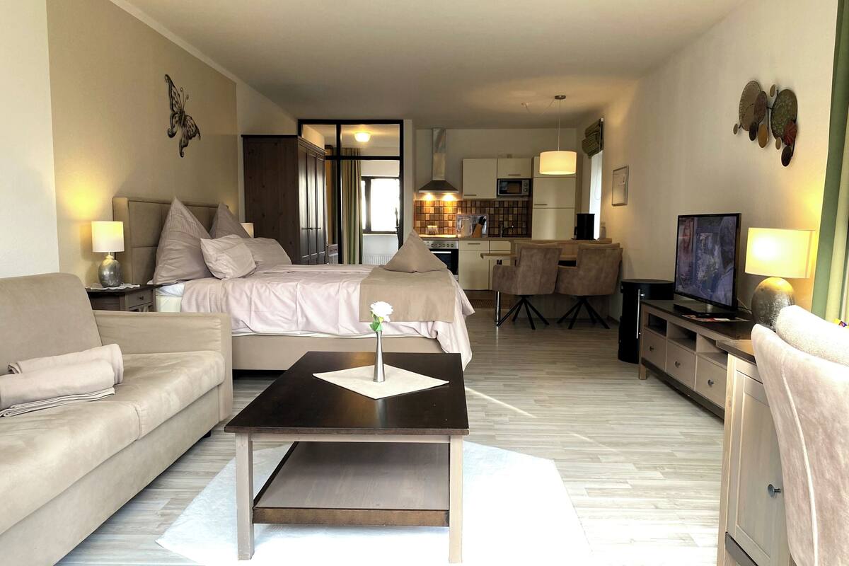 Une chambre d’hôtel moderne avec un lit, un canapé, une table basse, une télévision et, au fond, une petite cuisine.