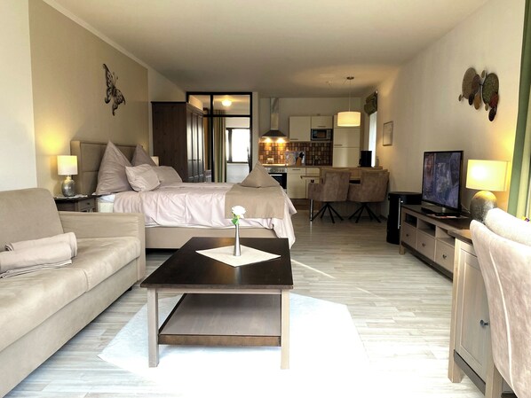 Habitación de hotel moderna con cama, sofá, mesa de centro, televisión y una kitchenette al fondo.