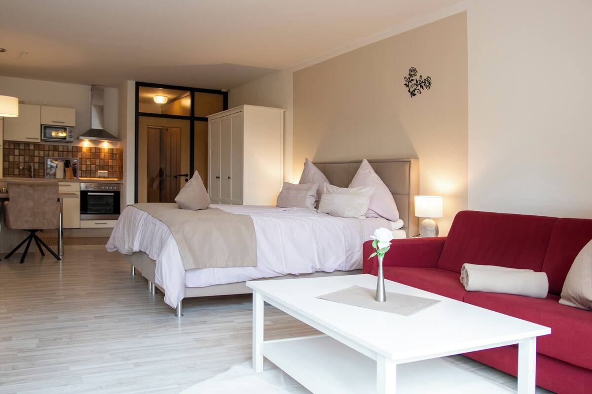 Une chambre avec un lit, un canapé rouge, une table basse blanche et, au fond, une petite cuisine.