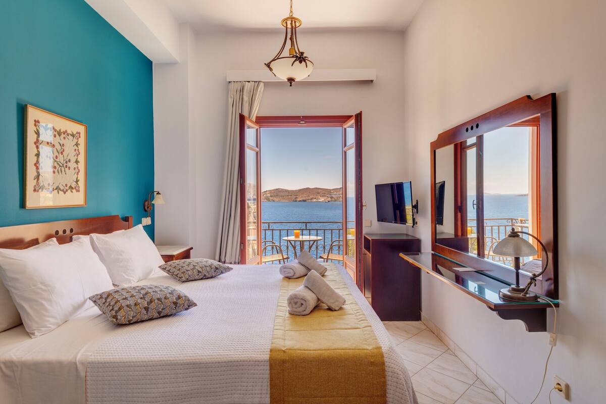 A hotel room with a bed, a TV, a desk, and a balcony with a view of the sea.