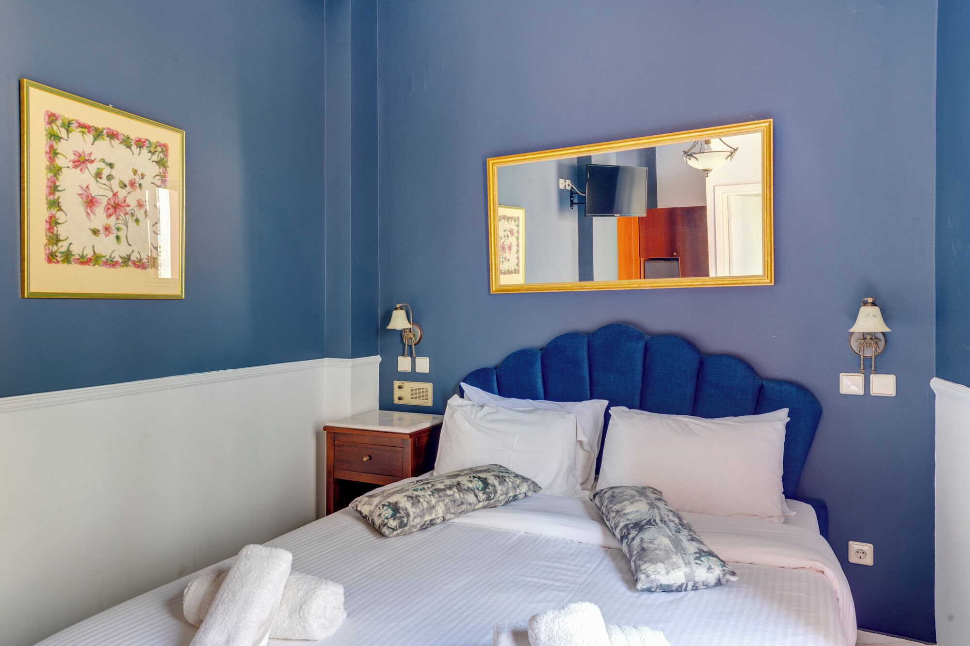 Una cama bien hecha con sábanas blancas y almohadas decorativas, un cabecero azul, una mesita de noche con una lámpara y una obra de arte floral enmarcada en la pared.