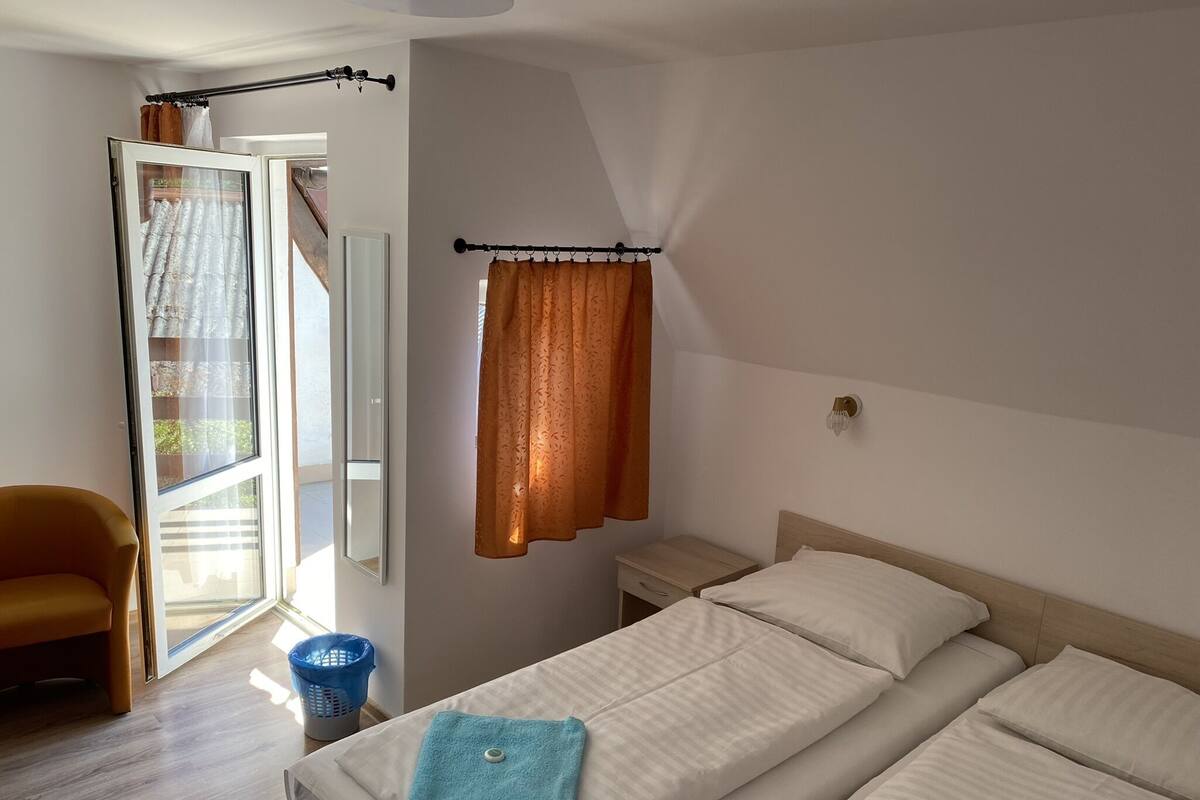 A bedroom with a bed, a chair, a nightstand, a window with curtains, and a view of the outside.