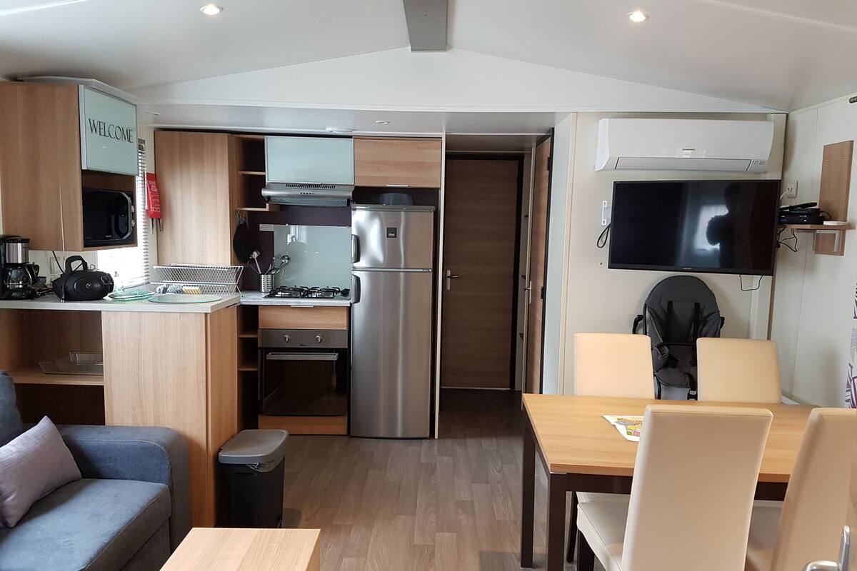 A compact kitchenette with a microwave, stove, and refrigerator, a sofa, a dining table with chairs, and a flat-screen TV mounted on the wall.