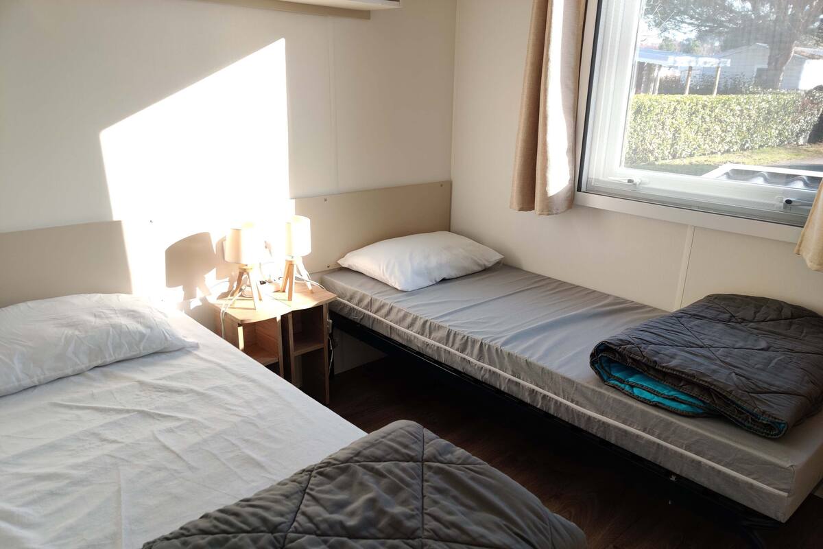 A small, compact room with two single beds, a desk with a lamp, and a window with curtains.