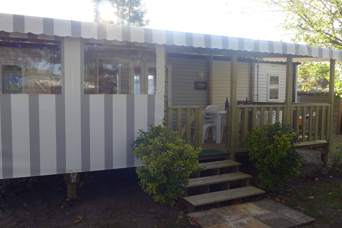 A small, single-story mobile home with a striped awning, a porch with a wooden railing, and a small set of steps leading to the entrance.