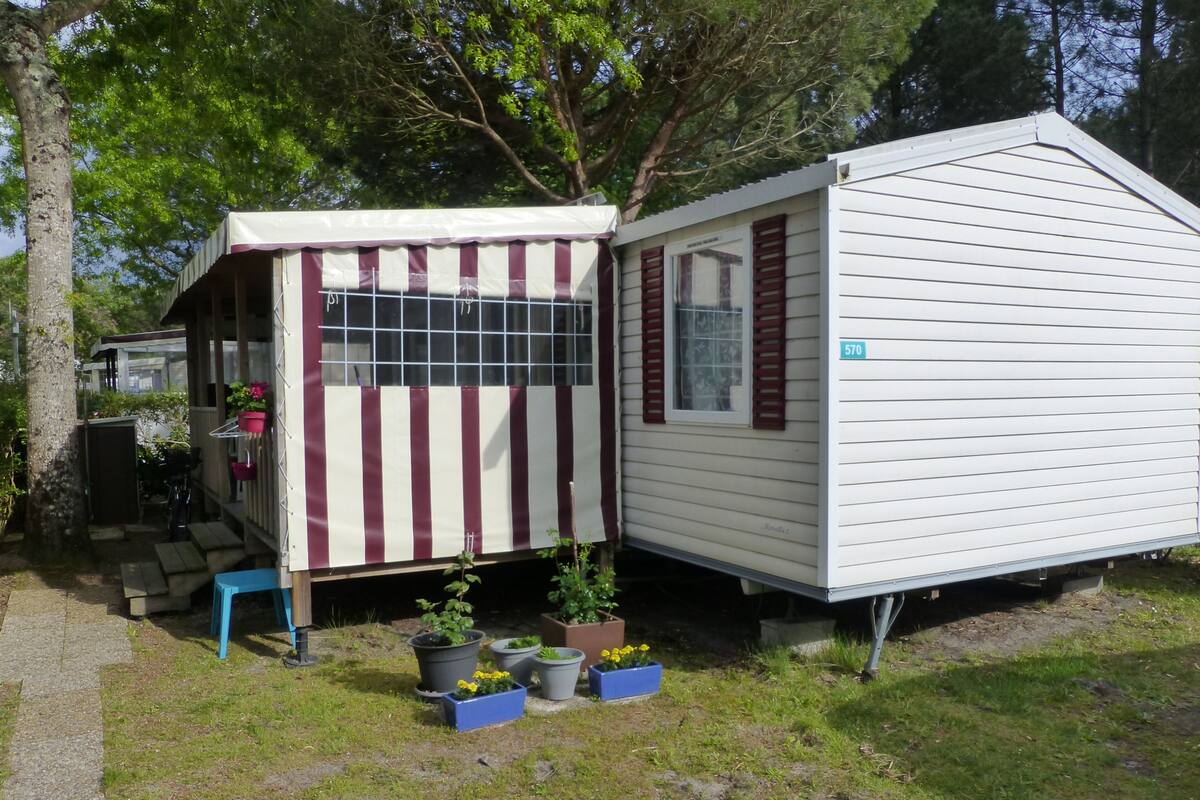 A mobile home with a striped awning and a window with shutters. There are potted plants and a blue chair outside.