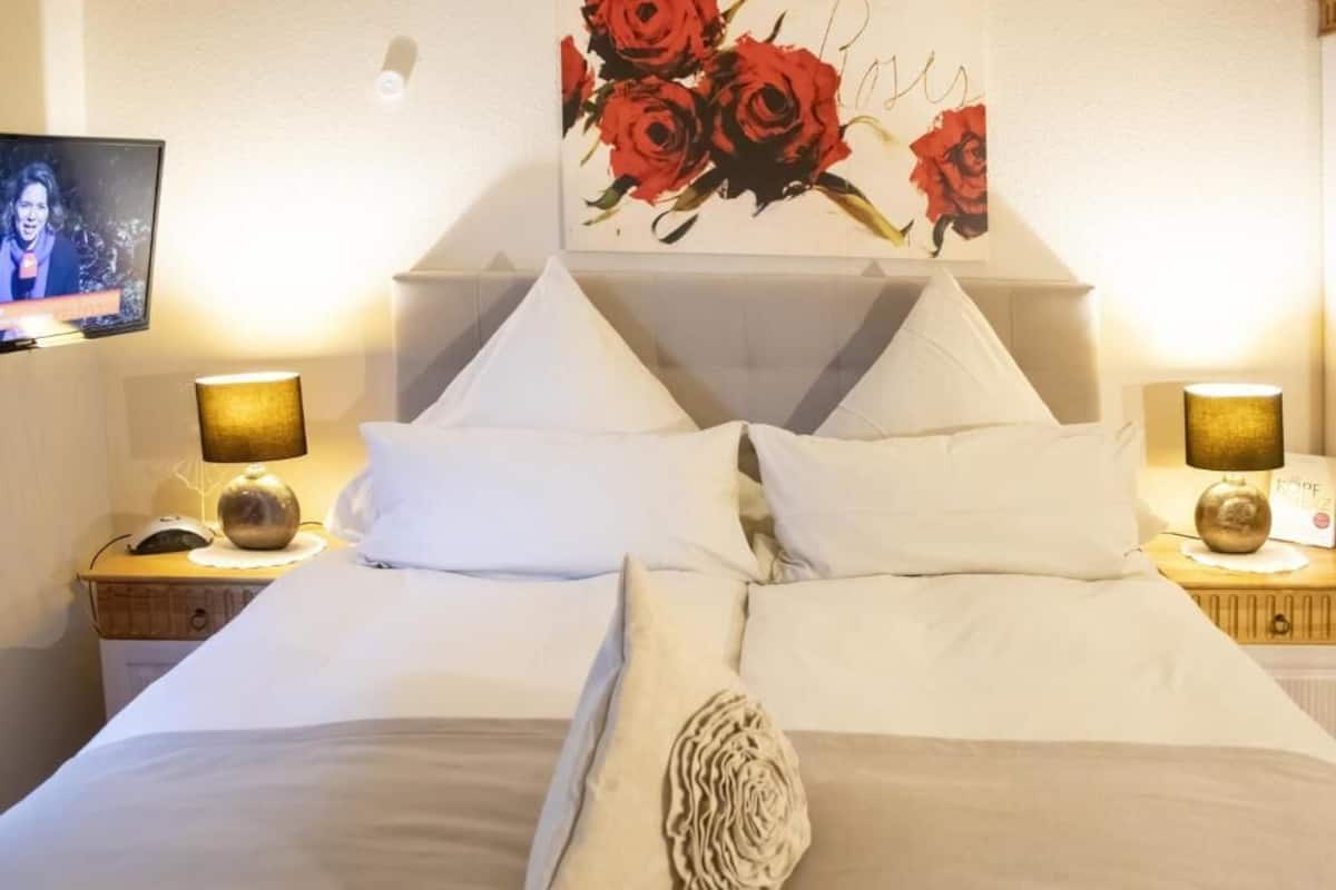 A neatly made bed with white linens, a decorative pillow, and a wall-mounted painting of red roses.