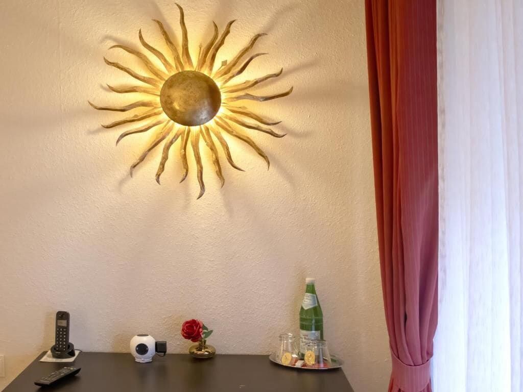 Un adorno de sol en la pared, un teléfono, una cámara, un jarrón con una flor y un vaso con una botella.