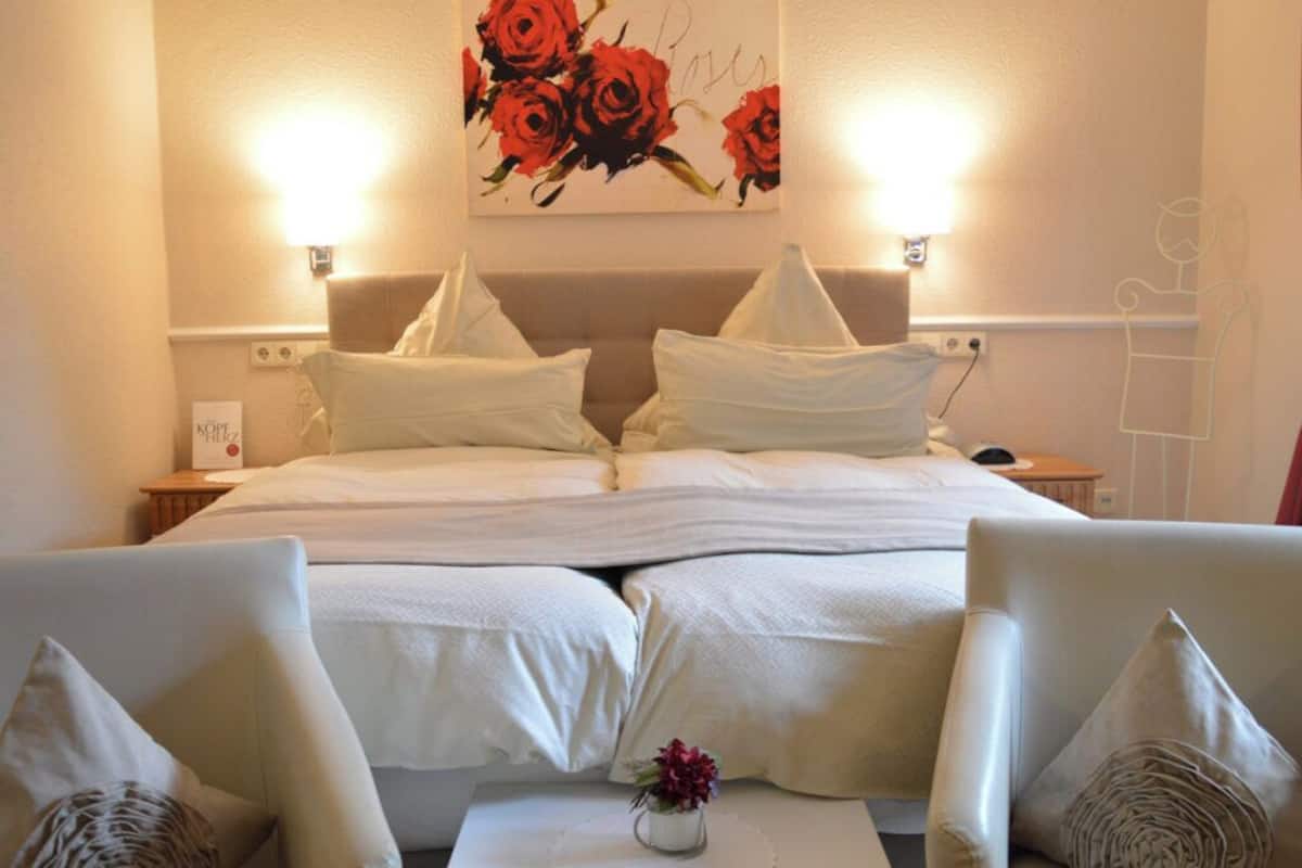 A hotel room with a bed, two bedside tables, a nightstand, a wall-mounted lamp, a painting of roses, and two armchairs with a small table between them.