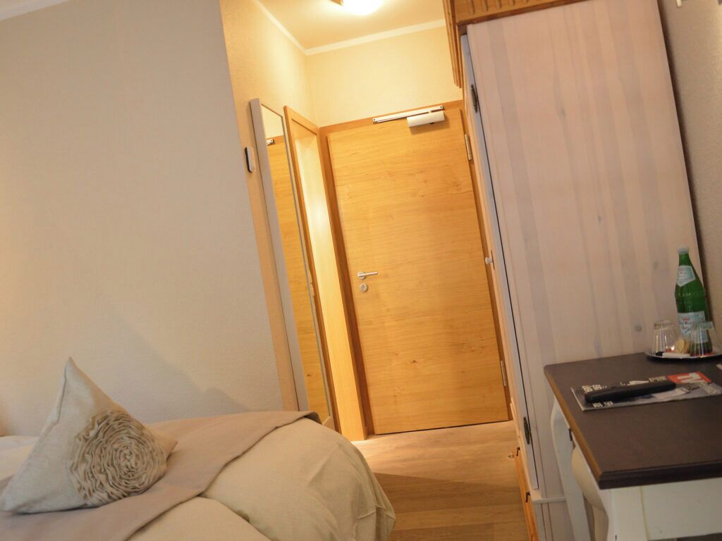 Una habitación de hotel con cama, una puerta de madera, un escritorio con una botella y un espejo.