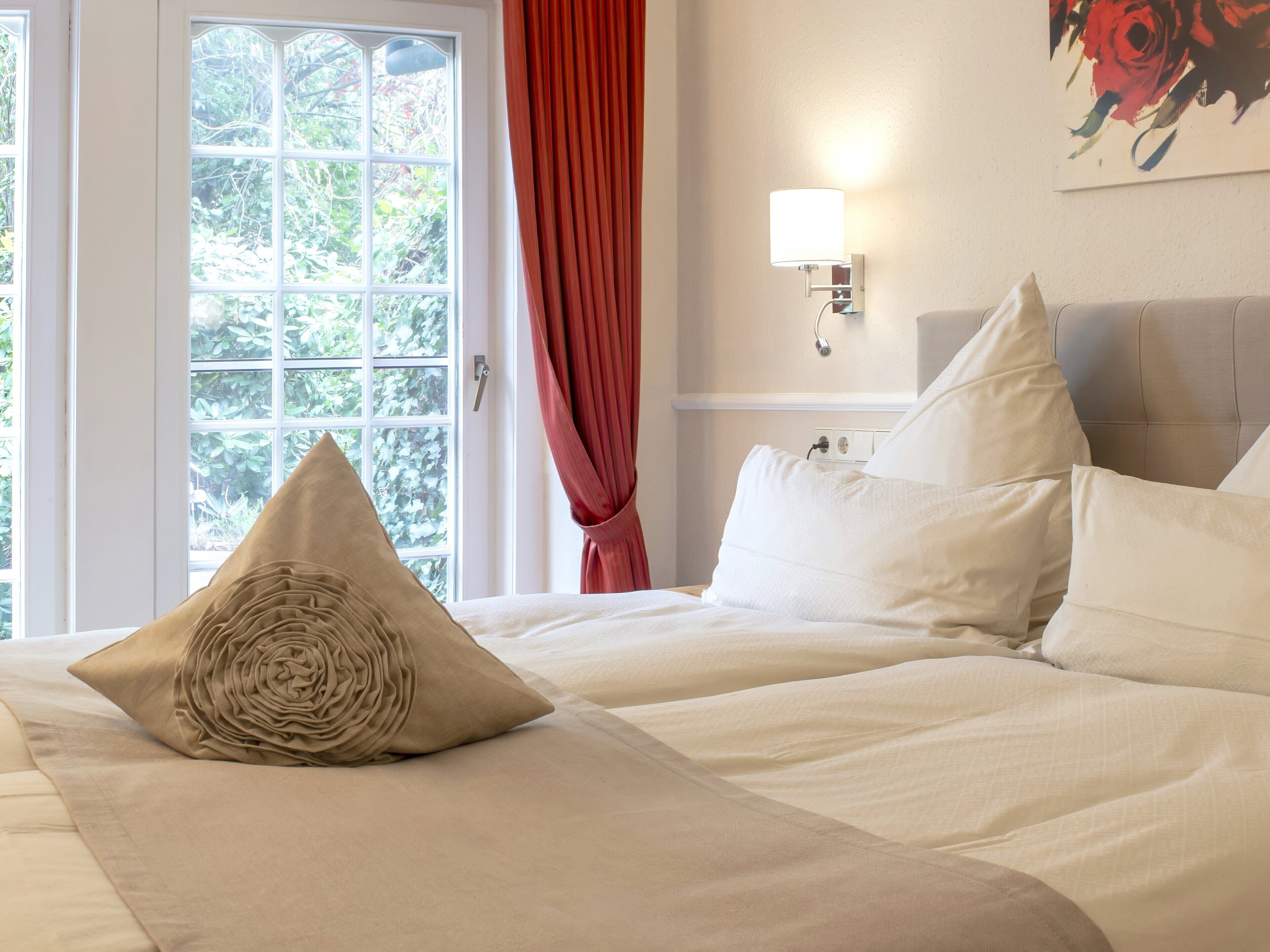 A hotel room with a bed, a window with curtains, a wall-mounted lamp, and a painting.