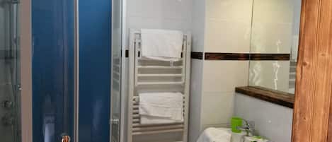 Un baño con cabina de ducha de vidrio, un perchero blanco con toallas, un lavamanos con un vaso verde y un espejo con marco de madera.
