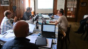 Un groupe de personnes se trouve dans une salle de réunion aux murs en bois, avec un grand écran et des ordinateurs portables posés sur une table.