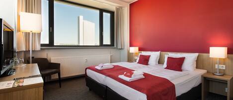 A hotel room with a large bed, two bedside tables with lamps, a desk with a computer and a chair, a window with a view of buildings, and a red accent wall.