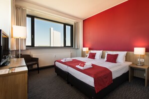 A hotel room with a large bed, two bedside tables with lamps, a desk with a computer and a chair, a window with a view of buildings, and a red accent wall. - BIENDO Hotel Chemnitz (Chemnitz)