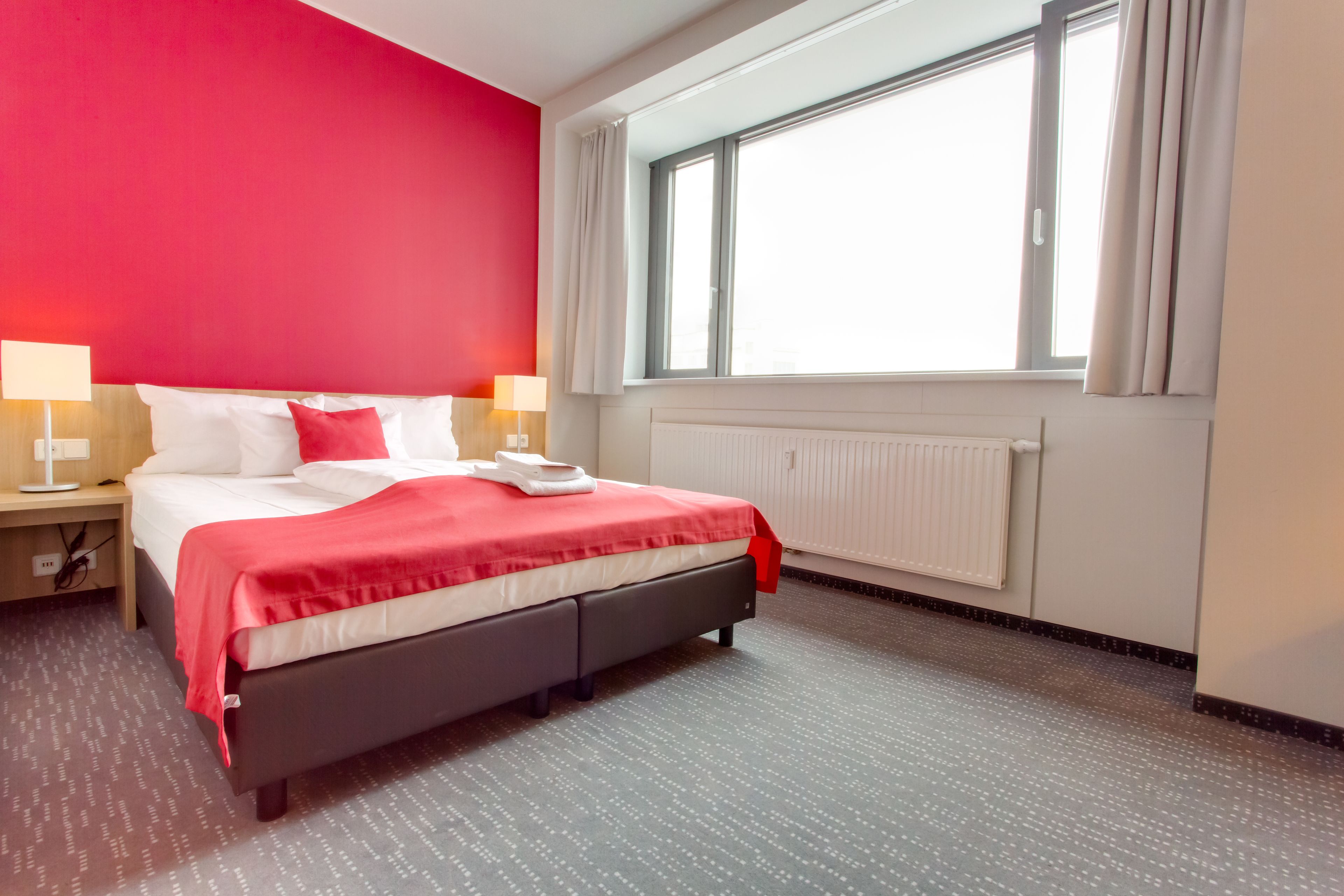A hotel room with a bed, two bedside lamps, a window with curtains, and a radiator.