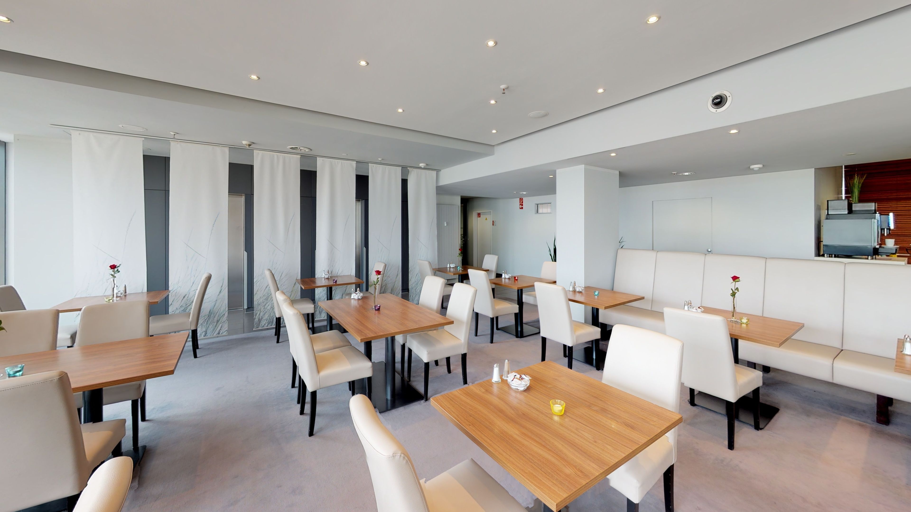 A modern dining area with multiple tables and chairs, a seating area with a bench, and a kitchen visible in the background.