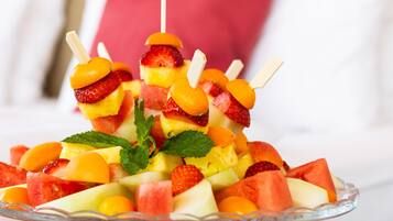 A fruit salad on a glass stand with a bed and pillows in the background.