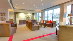 Un moderno vestíbulo de hotel con varias zonas de estar, un mostrador de recepción y grandes ventanales con vistas a la ciudad.