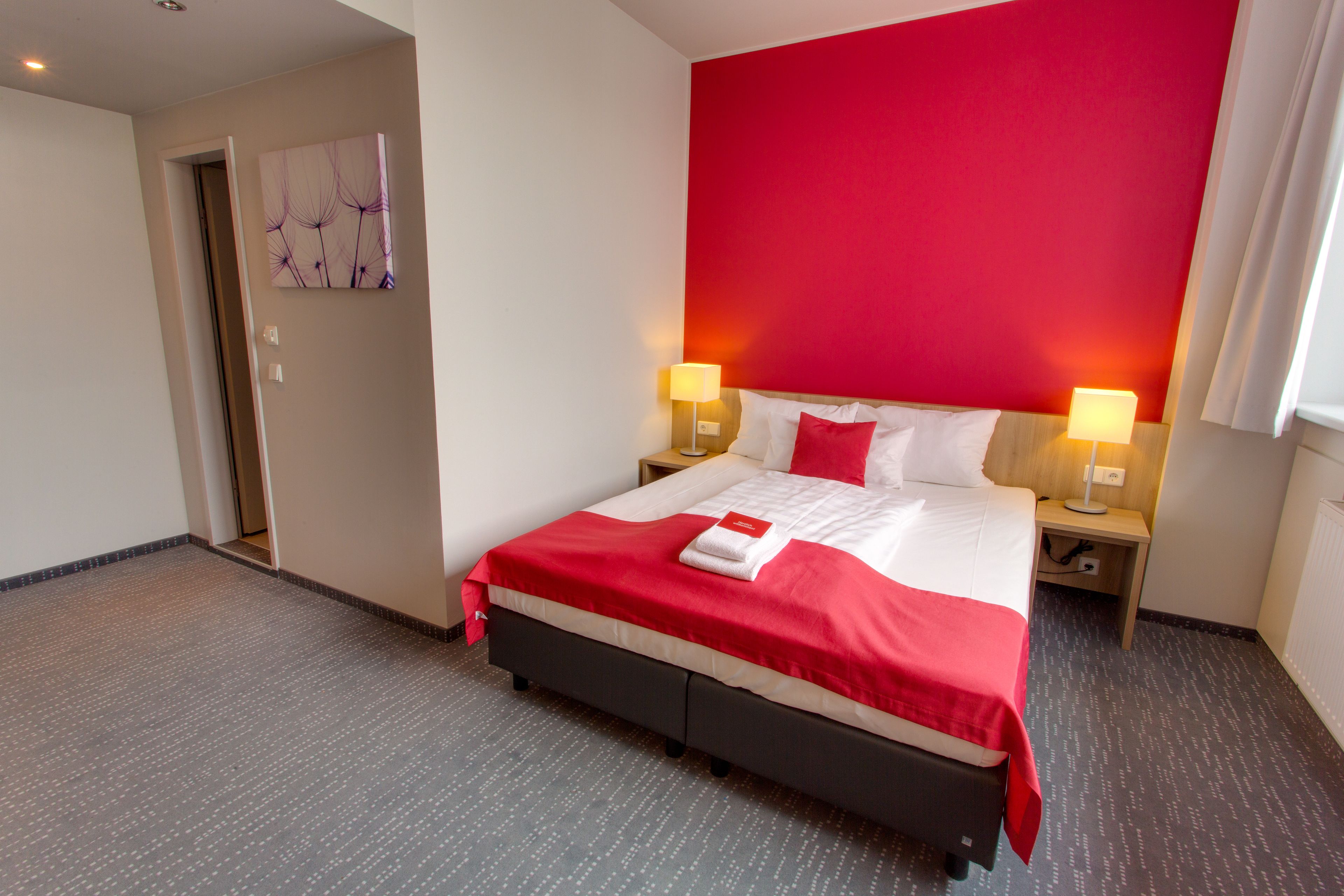 Una habitación de hotel con una cama grande, dos lámparas de noche, una pared de acento roja y una puerta que da a otra habitación.